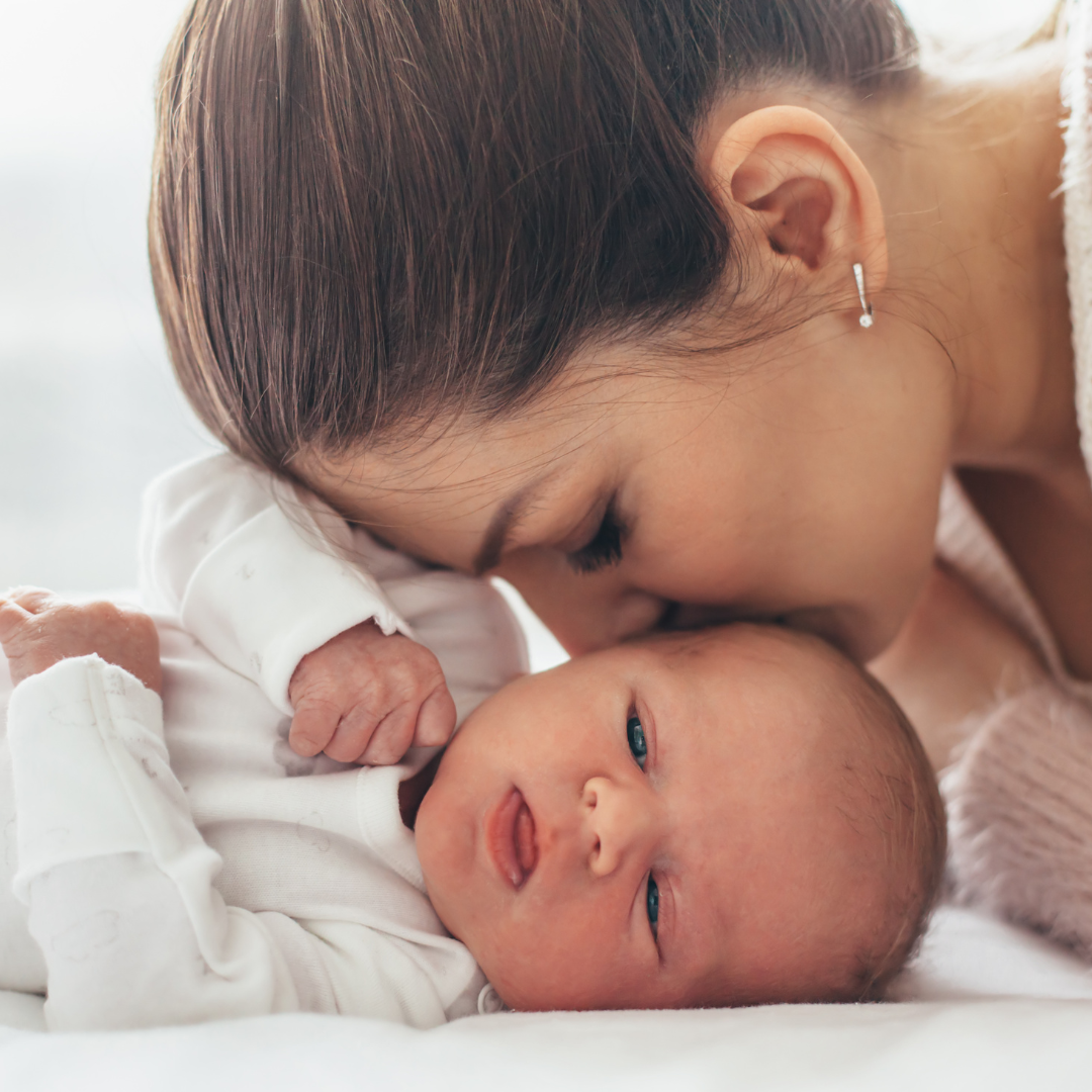neugeborenes Baby, das in seinem weißen Body liegt und von seiner Mutter auf die Wange geküsst wird 